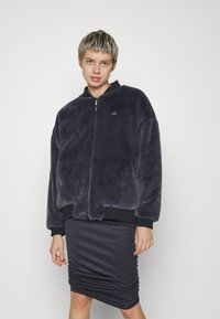 Jacheta bomber din blană artificială de culoare albastru marin, cu guler și manșete ribbate negre, având fermoar frontal și un branding subtil pe piept. Asortată cu o fustă mulată.