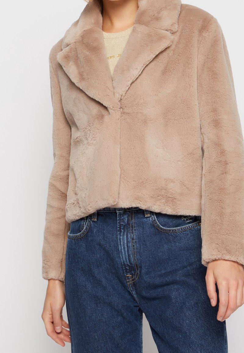 Veste en fausse fourrure beige à large col portée sur un haut clair, associée à un jean en denim bleu taille haute.