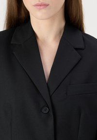Svart blazer laget av strukturert stoff, med definerte krager, enkeltknappingslukking og en brystlomme på venstre side.