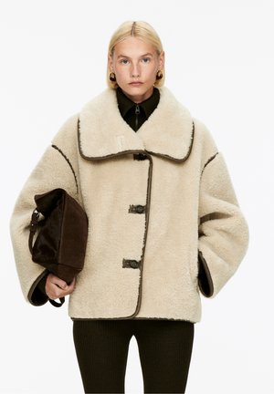 Beige Kunstfellmantel mit großem Kragen, braunen Akzenten und drei Knopfverschlüssen. Das Model hält eine dunkelbraune Handtasche.