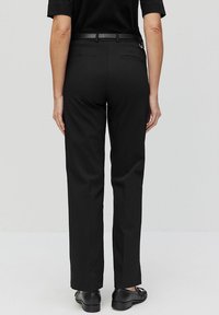 Zwarte broek met een rechtlijnig ontwerp, subtiele textuur, riemlussen en twee achterzakken. Draagt met zwarte schoenen, gezien van achteren.