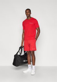 Κόκκινο βαμβακερό μπλουζάκι και σορτς με κεντημένο λογότυπο. Το μοντέλο κρατάει μαύρη ντουφελοθήκη, φοράει λευκά αθλητικά παπούτσια και κάλτσες με λογότυπο.