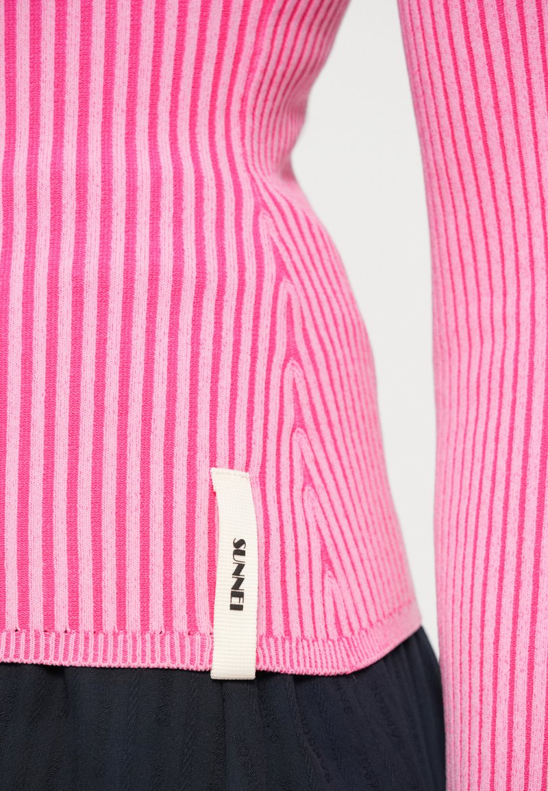 Fitted pink ribbed sweater with alternating lighter and darker stripes, featuring a woven label at the hem that reads "SUNNEI."