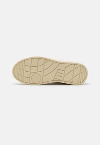 Suela de zapatilla de un beige claro con un patrón de banda de rodadura texturizado, que presenta un acabado suave y bordes redondeados. Detalle del logo visible en el lateral.