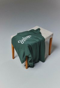 T-shirt en coton vert drapé sur un tabouret de couleur claire, avec un motif en texte blanc sur le devant. Texture douce, coupe décontractée.