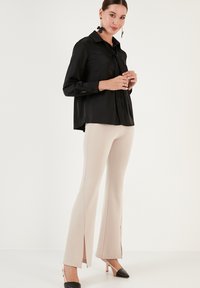 Bluză din satin negru cu detalii de legare, mâneci lungi și manșete cu nasturi, asortată cu pantaloni bej evazați, cu fante laterale, și pantofi cu toc ascuțit negri.