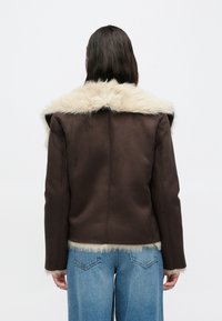 Καφέ μπουφάν από δερματίνη με μπεζ τεχνητή γούνα στον γιακά και το τελείωμα, με δομημένο σχέδιο και μακριά μανίκια. Φορεμένο με ανοιχτόχρωμα τζιν παντελόνια.