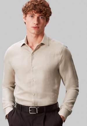 Ung mand med krøllet hår iført en beige skjorte med knapper og lange ærmer samt sort bælte, hænderne i lommerne, foran en ensfarvet grå baggrund.