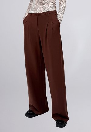 Osoba nosi ciemnobrązowe spodnie z wysokim stanem i szerokimi nogawkami, z przodu z zakładkami i kieszeniami, czarne masywne buty oraz długi rękaw w panterkę.