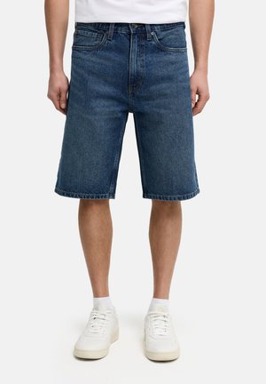 Hombre con pantalones cortos de mezclilla azul hasta la rodilla, calcetines blancos y zapatillas blancas, de pie frente a un fondo blanco liso.