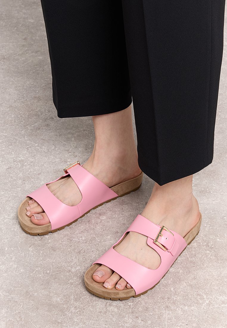 Sandales glissantes roses avec une finition mate, comportant une large sangle, un design découpé et un accent de boucle dorée. Semelle en liège pour plus de confort.