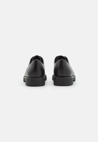 Chaussures en cuir noir avec des talons arrondis, texture lisse et design minimaliste. Elles présentent un profil bas et des coutures subtiles le long des bords.