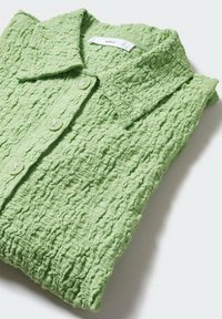 Chemise en tissu texturé vert clair avec col et trois boutons, soigneusement pliée sur une surface blanche.