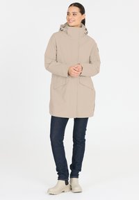 Veste imperméable beige avec un col haut, des poches latérales et une capuche. Portée avec un jean en denim foncé et des bottines de couleur claire.