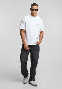Hvid bomulds t-shirt, løs pasform, med logo detalje. Sorte denim jeans med lige snit. Grå og hvide sneakers med striber. Sorte solbriller.