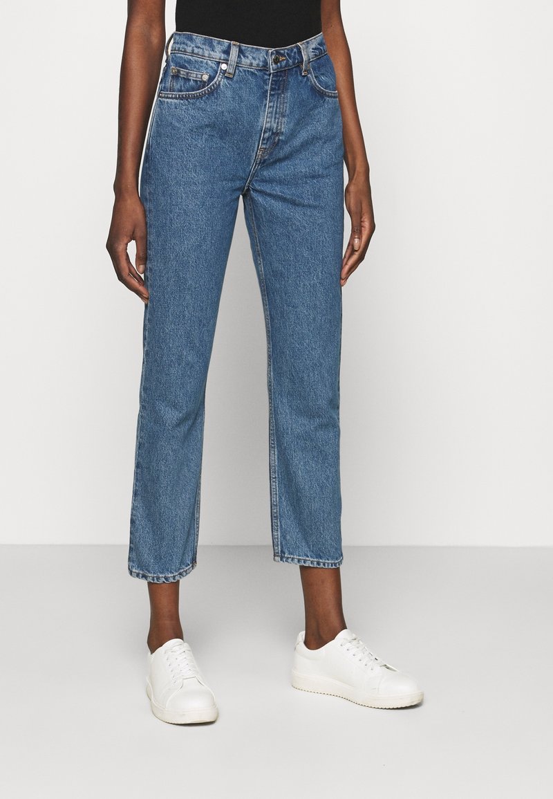 Personne portant un jean bleu taille haute à jambe droite associé à des baskets blanches, debout devant un fond clair uni.