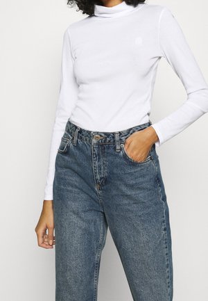 Vrouw met een witte, lange mouws coltrui, ingestopt in een high-waisted, loszittende blauwe spijkerbroek, met één hand in de zak.