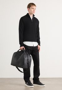 Melns zip-up džemperis virs baltas krekla, melni bikses un melni sporta apavi. Modelis tur melnu ādas sporta somu ar diviem rokturiem.