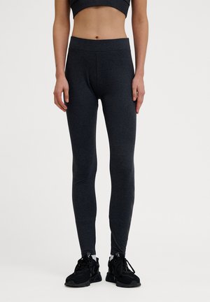 Kobieta ubrana w dopasowane ciemnoszare legginsy i czarne buty sportowe, stojąca na gładkim jasnym tle.