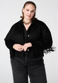 Zwarte suède cropped jas met franjes, zilverkleurige knopen en een losse pasvorm. Gecombineerd met donkere jeans voor een monochrome uitstraling.