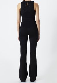 Kobieta stojąca tyłem, ubrana w bezrękawnik czarny z koronkowym tyłem oraz czarne spodnie w dzwony na białym tle.