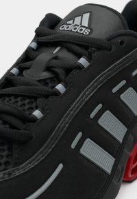Gros plan d'une basket Adidas noire avec des lacets noirs, des détails de bandes grises, une tige en mesh et un amorti rouge visible au talon.