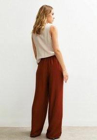 Top senza maniche beige abbinato a pantaloni larghi marrone ruggine con tessuto testurizzato e vita elasticizzata, mostrato da un angolo leggermente posteriore.