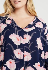 Blusa blu navy con motivo floreale rosa, scollo a V e chiusura a bottoni. Tessuto morbido con una vestibilità ampia e una leggera lucentezza.