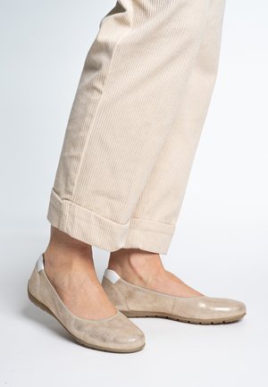 Beige flats made of textured material, featuring a rounded toe and white accents on the heel, worn with rolled-up cream corduroy pants.