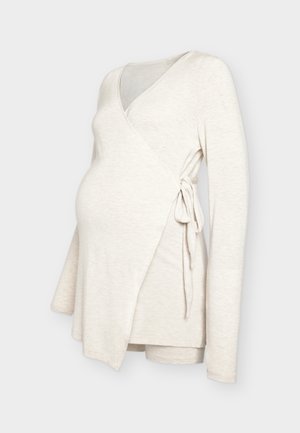 Haut de maternité beige à manches longues avec nouage latéral, conçu pour s’adapter à un ventre qui s’arrondit, présenté sur un fond neutre.