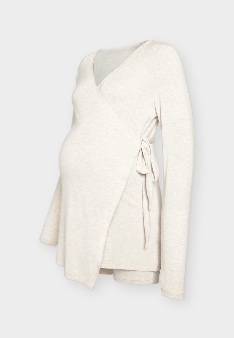 Haut de maternité beige à manches longues avec nouage latéral, conçu pour s’adapter à un ventre qui s’arrondit, présenté sur un fond neutre.