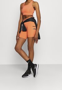 Top deportivo naranja y pantalones cortos, con una chaqueta negra atada a la cintura. Zapatillas de correr negras con detalles blancos y calcetines.