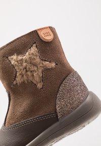 Hnědé semišové boty s chlupatým hvězdovým detailem, hnědým koženým ochranným krytem na patě a třpytivým hnědým akcentem na podrážce s logem.