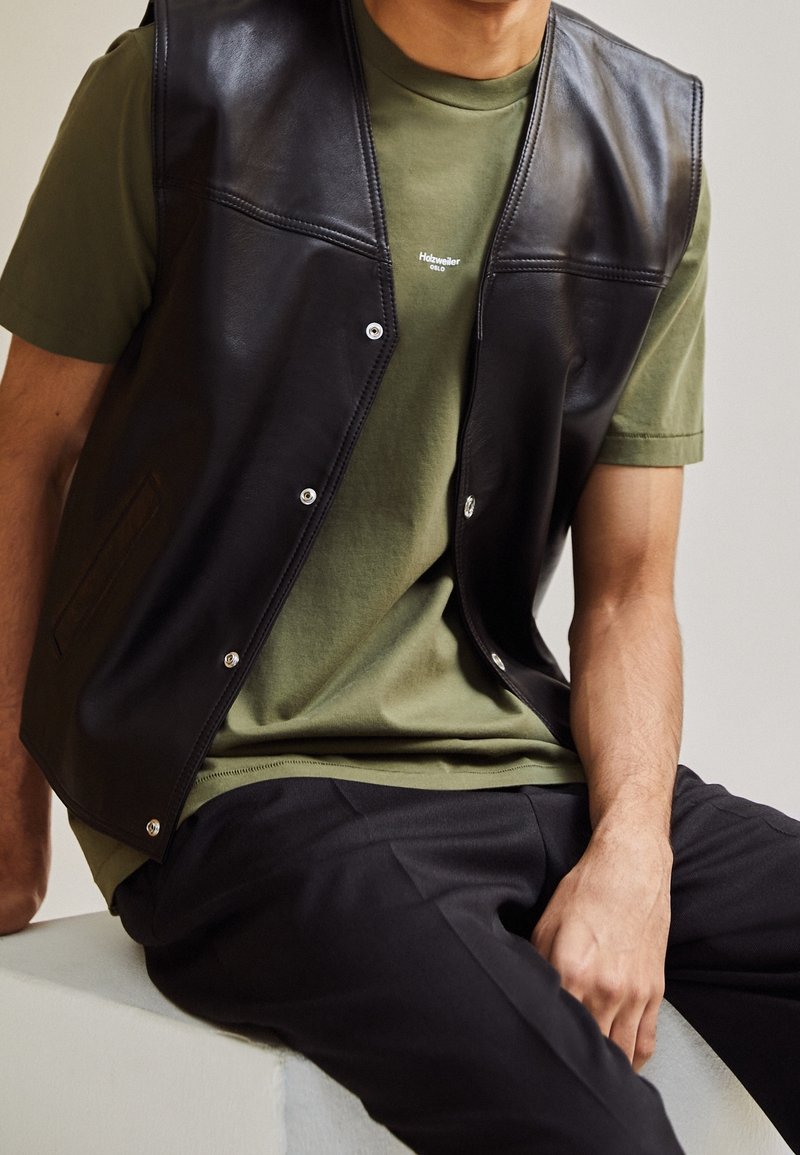 Homme assis portant un gilet en cuir noir avec des boutons-pression sur un t-shirt vert olive et un pantalon noir, les mains reposant sur les genoux et la surface.