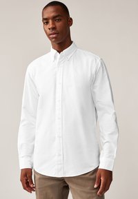 Weißes Baumwollhemd mit durchgehender Knopfleiste, ausgestelltem Kragen, langen Ärmeln und abgerundetem Saum. Glatte Textur mit dezentem Markenlogo.
