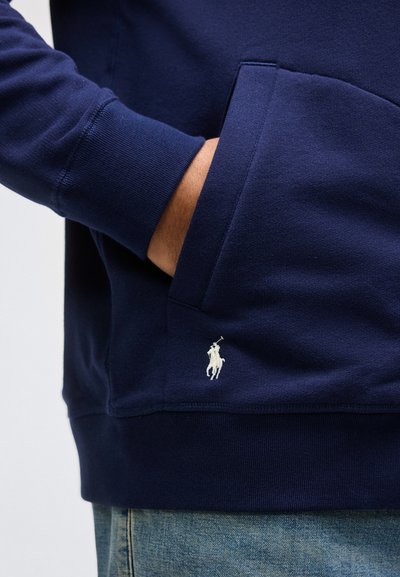 Χέρι στην τσέπη μπλε σκούρου φούτερ με λευκό κεντημένο λογότυπο παίκτη πόλο πάνω από μπλε τζιν.