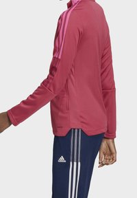 Chaqueta deportiva rosa con cuello corto de pie, mangas largas y acentos rayados rosas en los hombros. Combinada con pantalones marinos que presentan tres rayas.