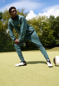 Tuta sportivo verde con giacca con zip e pantaloni, caratterizzato da accenti gialli. Scarpe da calcio nere e bianche su un campo verde.