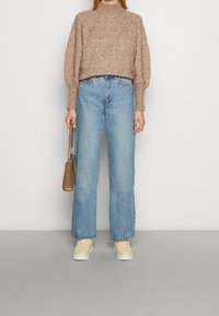 Pull en tricot marron clair avec un col haut, associé à un jean bleu à jambes larges et des baskets jaunes. Sac bandoulière marron à la main.