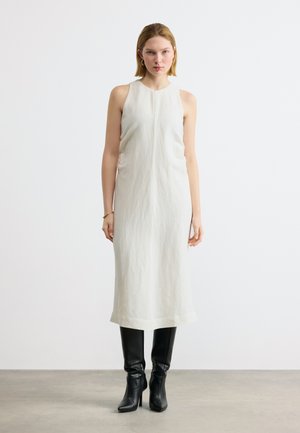 Nő áll, ujjatlan fehér midi ruhát és fekete, térdig érő, magas sarkú csizmát visel, egyszínű világos háttér előtt.