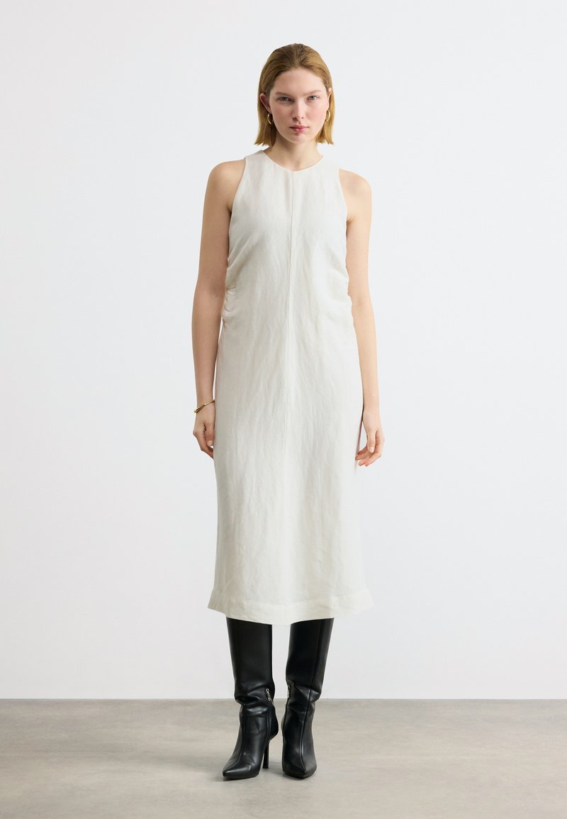 Γυναίκα που στέκεται, φορώντας ένα λευκό αμάνικο μίντι φόρεμα και μαύρες μπότες με τακούνι έως το γόνατο, μπροστά σε ένα απλό φωτεινό φόντο.