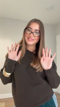 Rjavo pulover z rebrastimi manšetami, prikazuje dolge lase in zlato zapestnico. Roke dvignjene z razprtim prsti, mehka osvetlitev na nevtralnem ozadju.