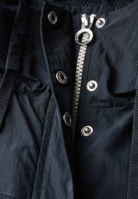 Marineblå jacka med silverdragkedja, tryckknappar och texturerad tyg. Detaljer inkluderar en flätad dragsko och ett fodrat krage.