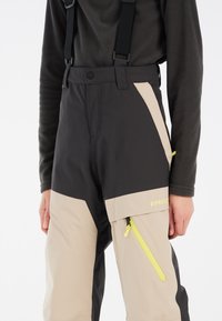Pantalon de ski résistant à l'eau avec bretelles réglables. Présente un schéma de couleurs contrasté beige et noir, avec une poche zippée et un détail de logo.