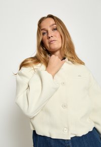 Witte gestructureerde jas met een knoopsluiting aan de voorkant en lange mouwen, met een borstzak en een relaxte pasvorm. Gecombineerd met donkerblauwe denim jeans.