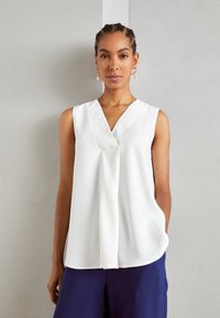 Canotta bianca senza maniche con scollo a V, fronte drappeggiato e texture morbida, abbinata a pantaloni blu navy in una vestibilità rilassata.