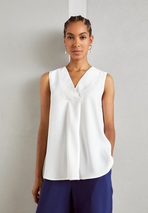 Blusa blanca sin mangas con escote en V, frente drapeado y textura suave, combinada con pantalones azul marino de corte relajado.