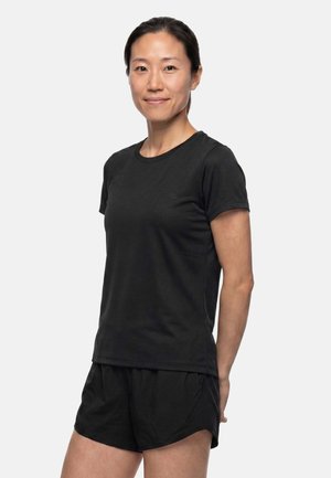 Femme debout face à l'avant, vêtue d'un t-shirt noir à manches courtes et d'un short noir, souriante avec les mains derrière le dos.