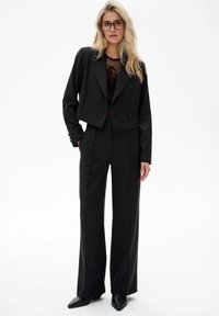 Zwarte op maat gemaakte cropped blazer met dubbele knopen, gecombineerd met hoge taille met brede pijpen. Model draagt een doorschijnende top eronder.