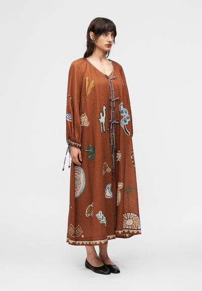 Γυναίκα που στέκεται, φορώντας φόρεμα μήκους μέχρι τους αστραγάλους σε σκουρόχρωμο σκουριασμένο χρώμα με μακριά μανίκια, μπροστινούς δεσμούς και πολύχρωμα αφηρημένα μοτίβα, και μαύρα παπούτσια μπαλαρίνες.